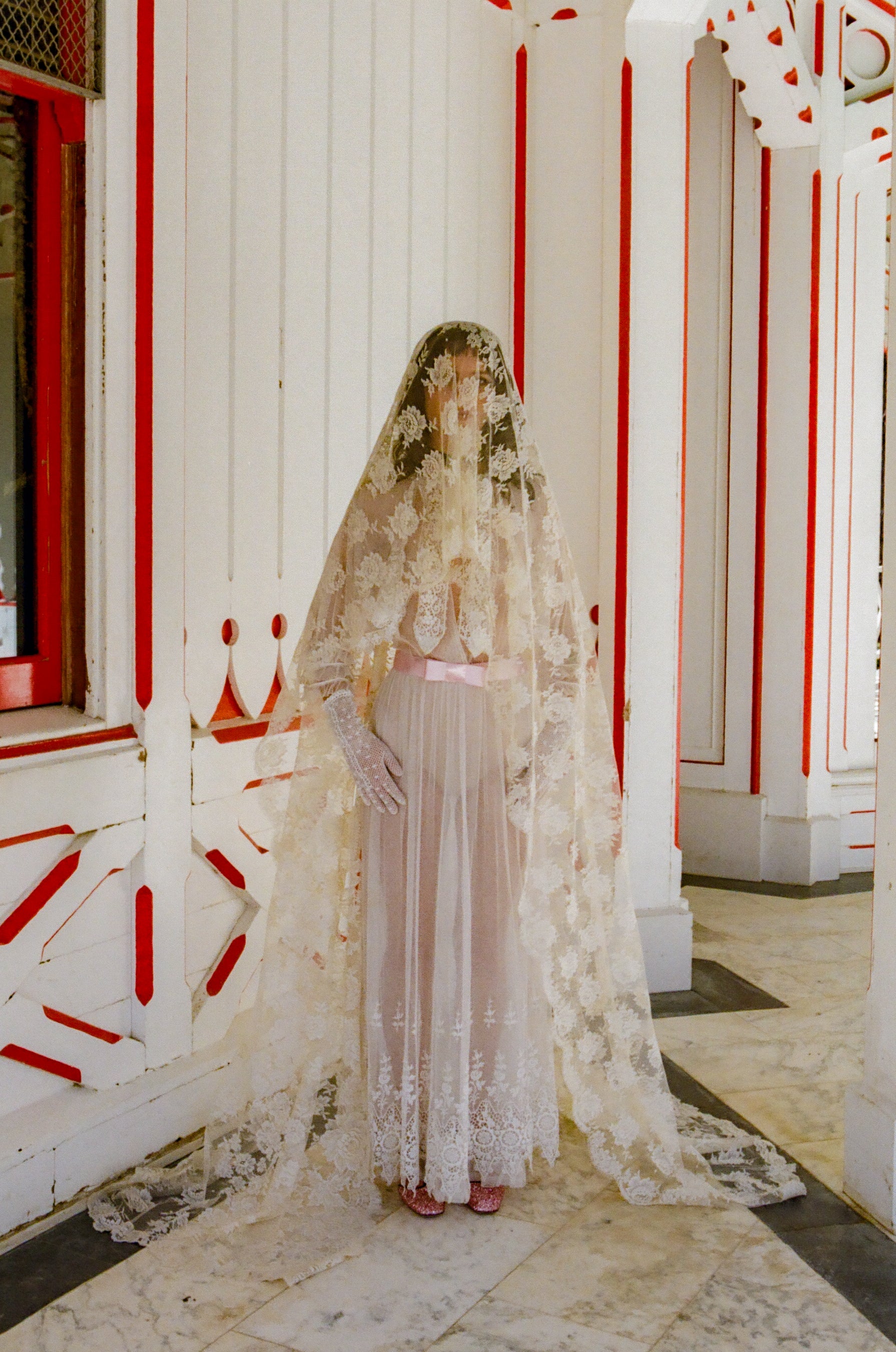Woman in a white lace dress standing in a decorated interior setting with red and white patterns.