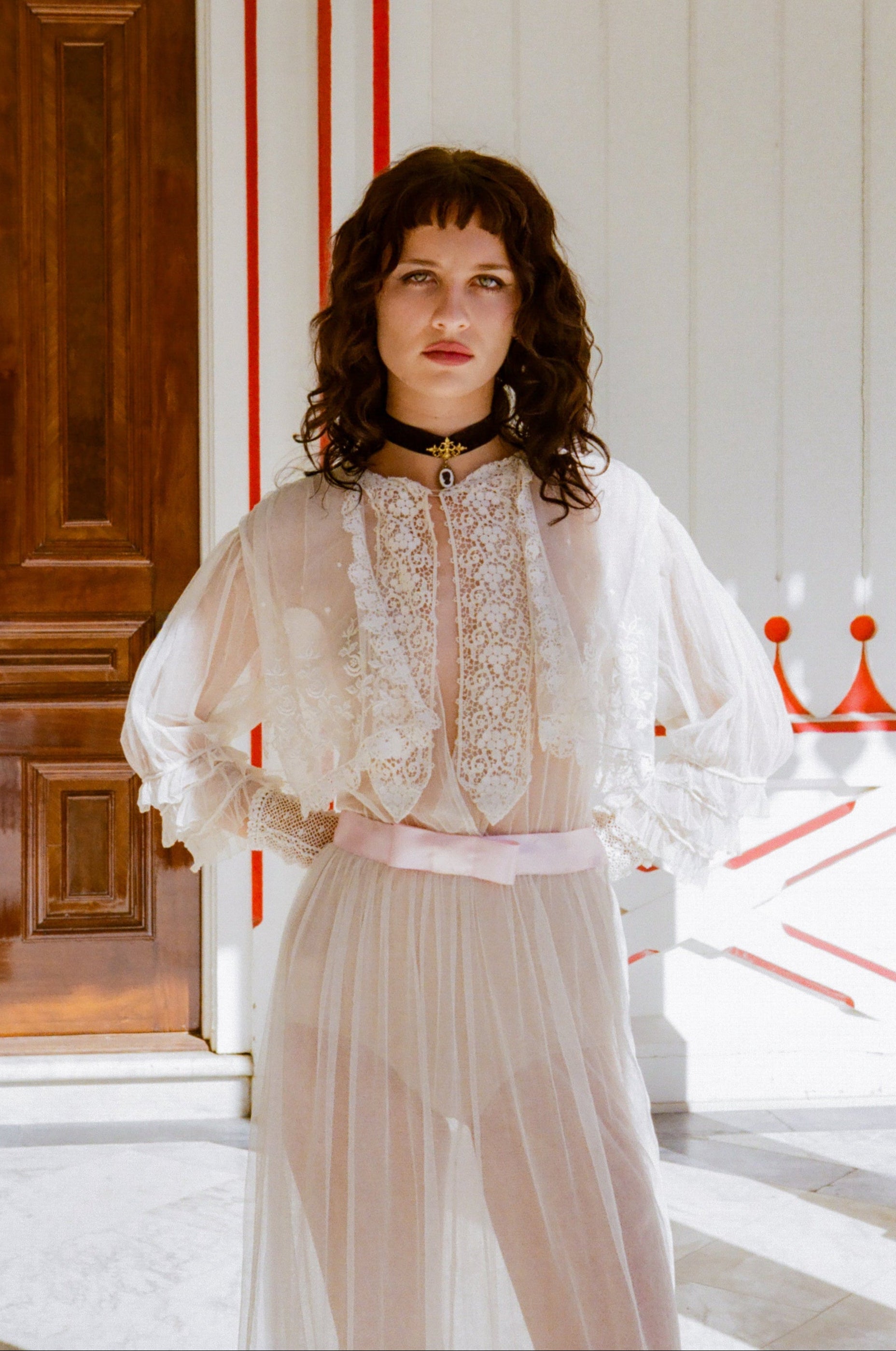 Woman wearing a white lace top and light-colored skirt in a room with wooden door and red accents.