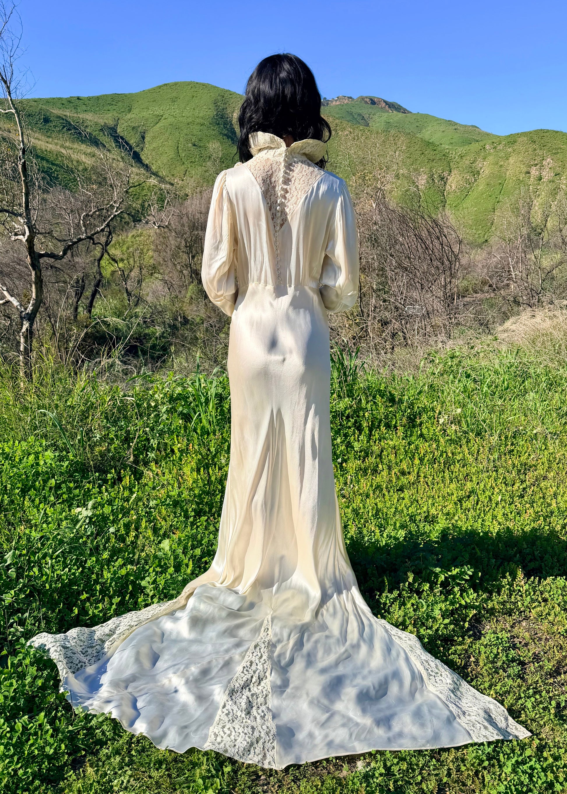 Person wearing an antique 1930's silk ivory wedding gown long, light-colored dress standing in a natural setting with green hills and blue sky.