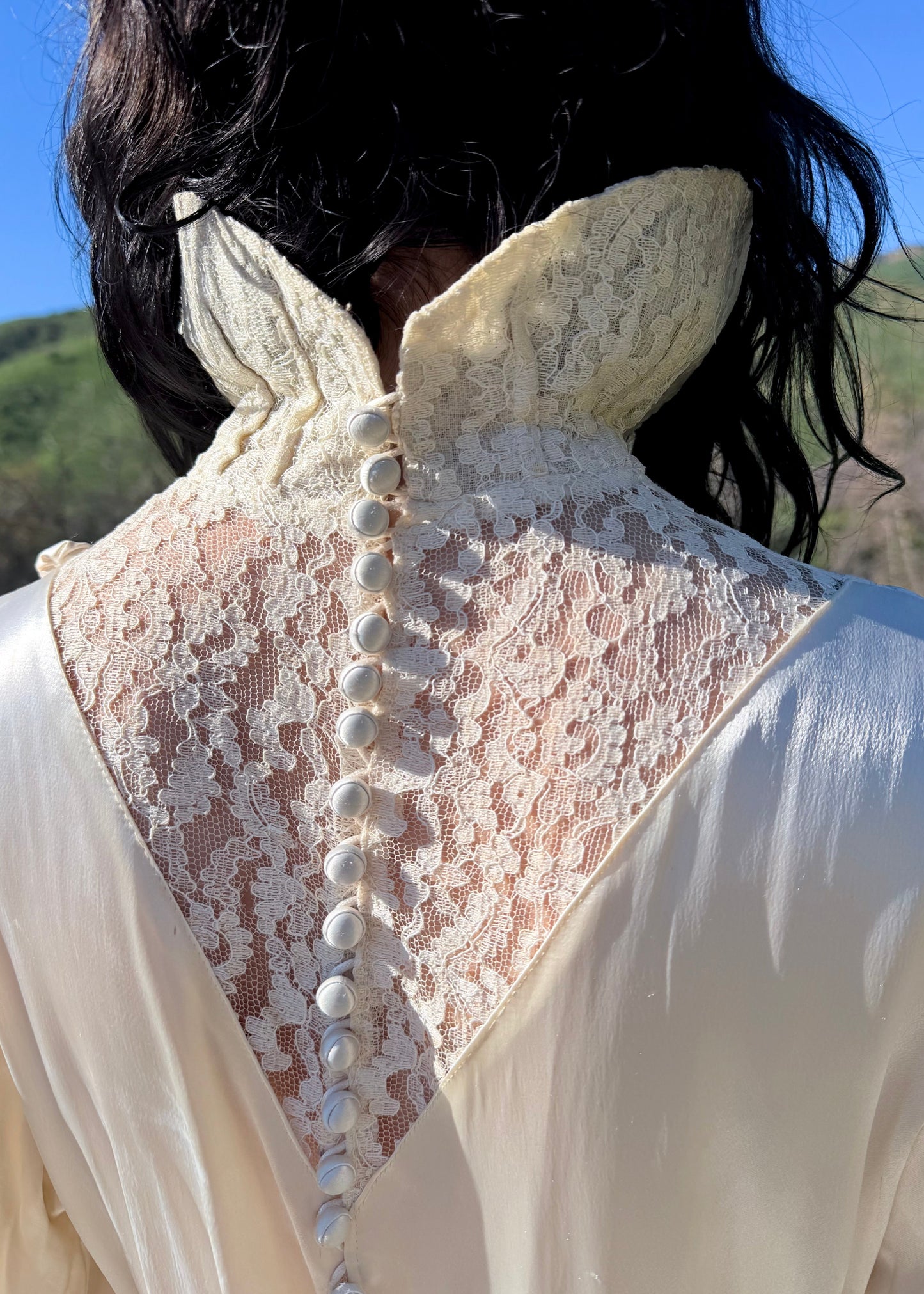 Person wearing an antique 1930's silk ivory wedding gown long, light-colored dress standing in a natural setting with green hills and blue sky.