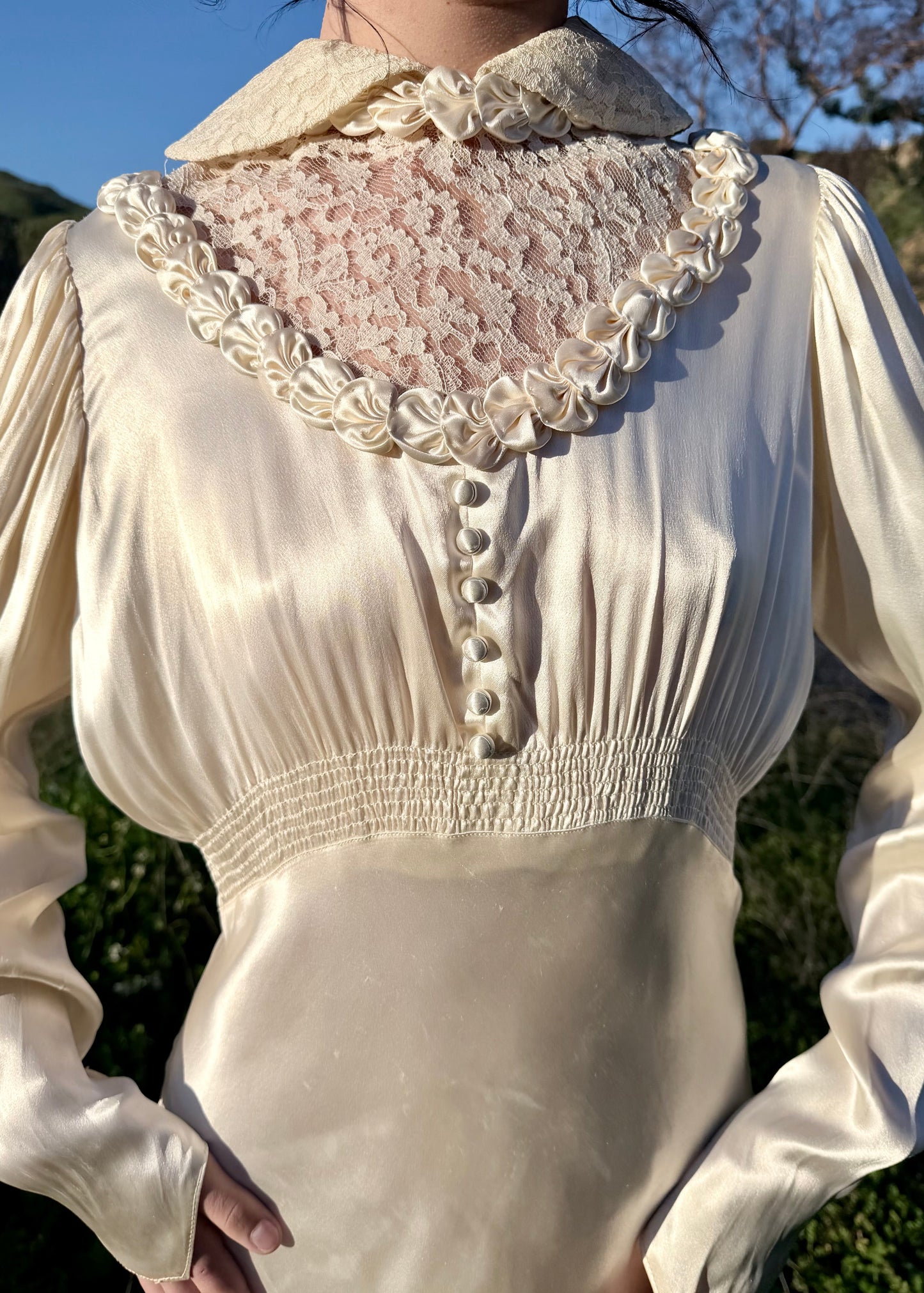 Person wearing an antique 1930's silk ivory wedding gown long, light-colored dress standing in a natural setting with green hills and blue sky.