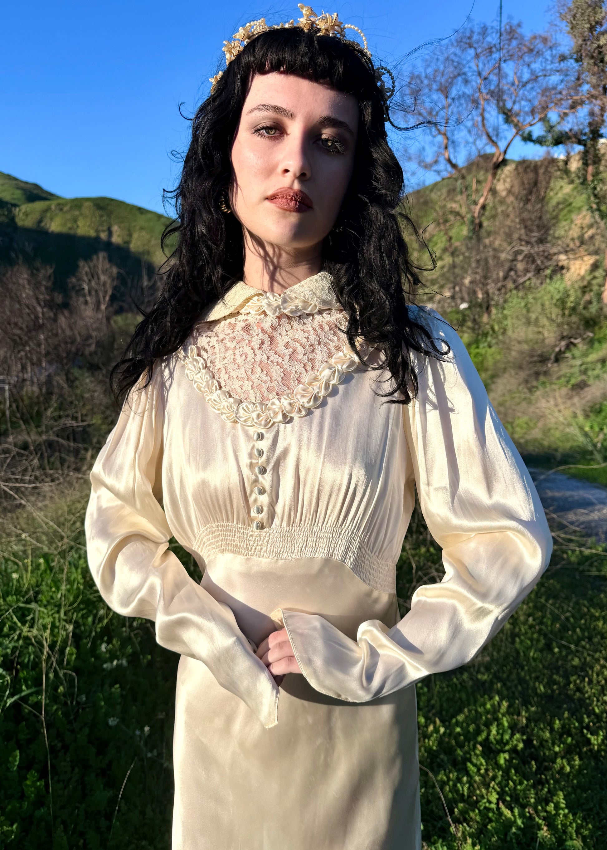 Person wearing an antique 1930's silk ivory wedding gown long, light-colored dress standing in a natural setting with green hills and blue sky.