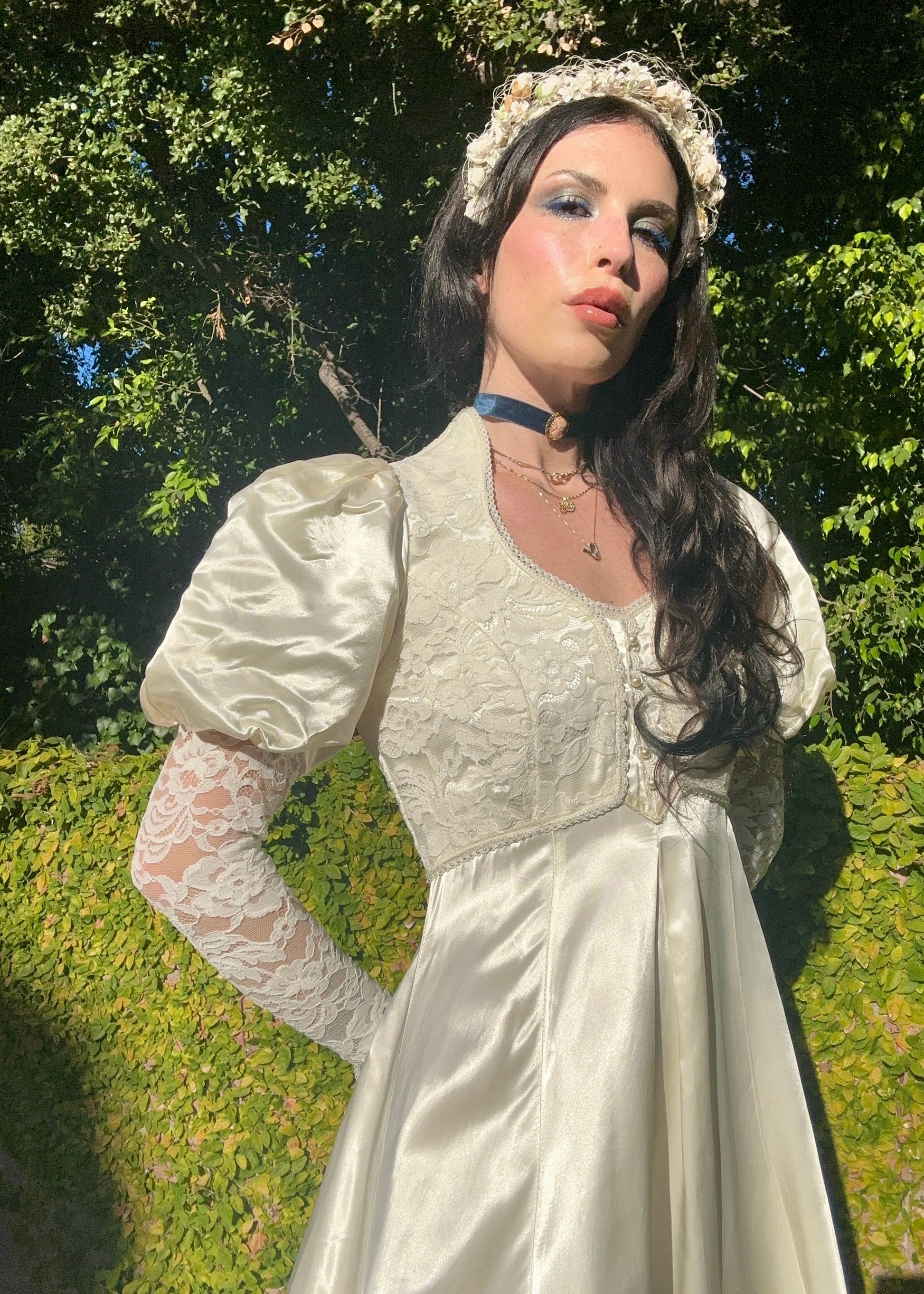 Woman in a white vintage dress with lace sleeves and a floral headpiece, standing outdoors with greenery.