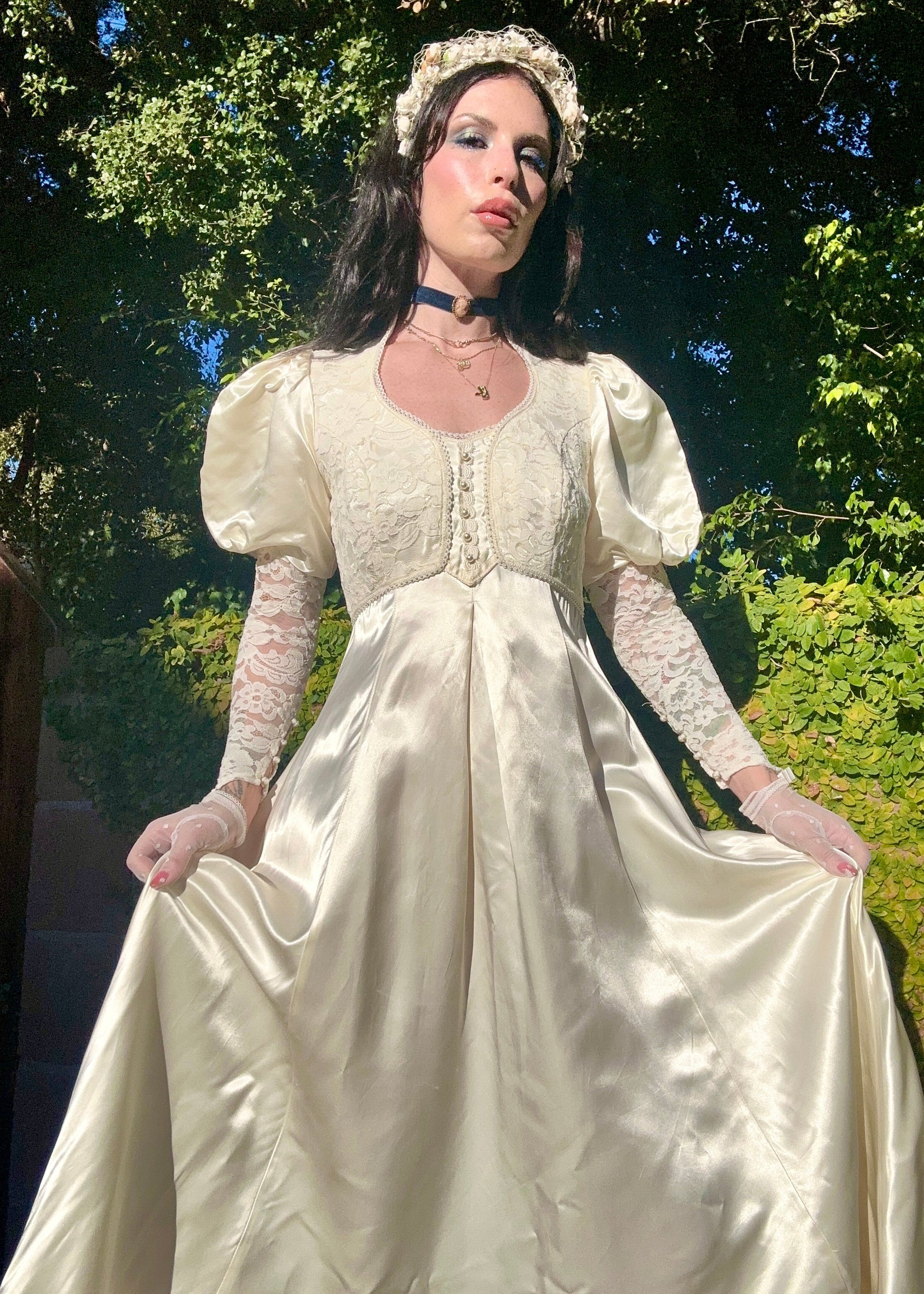 Woman in a white vintage dress with puffed sleeves and gloves standing outdoors with greenery in the background.
